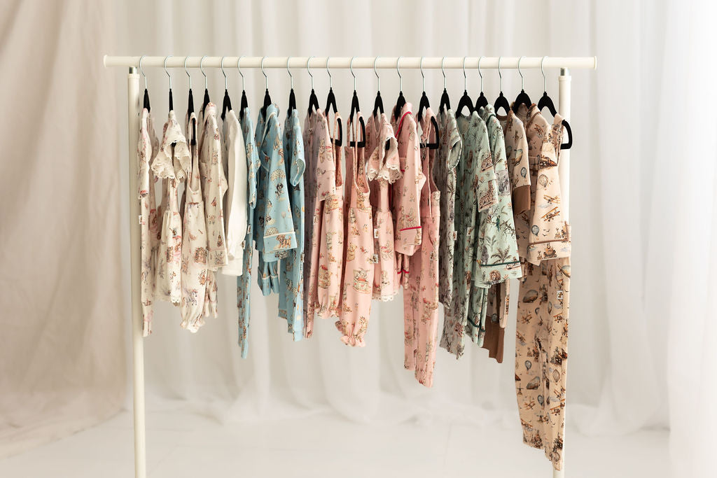 A white clothing rack holding multiple baby clothing sets in assorted patterned fabrics, photographed against a soft white draped backdrop.