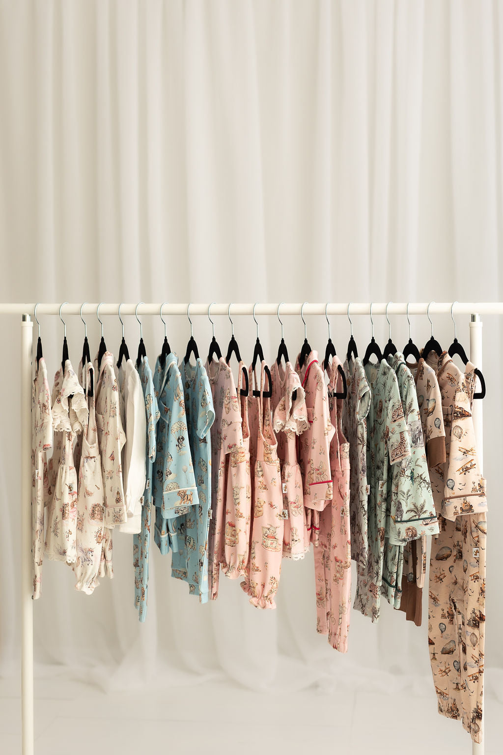 A white clothing rack holding multiple baby clothing sets in assorted patterned fabrics, photographed against a soft white draped backdrop.